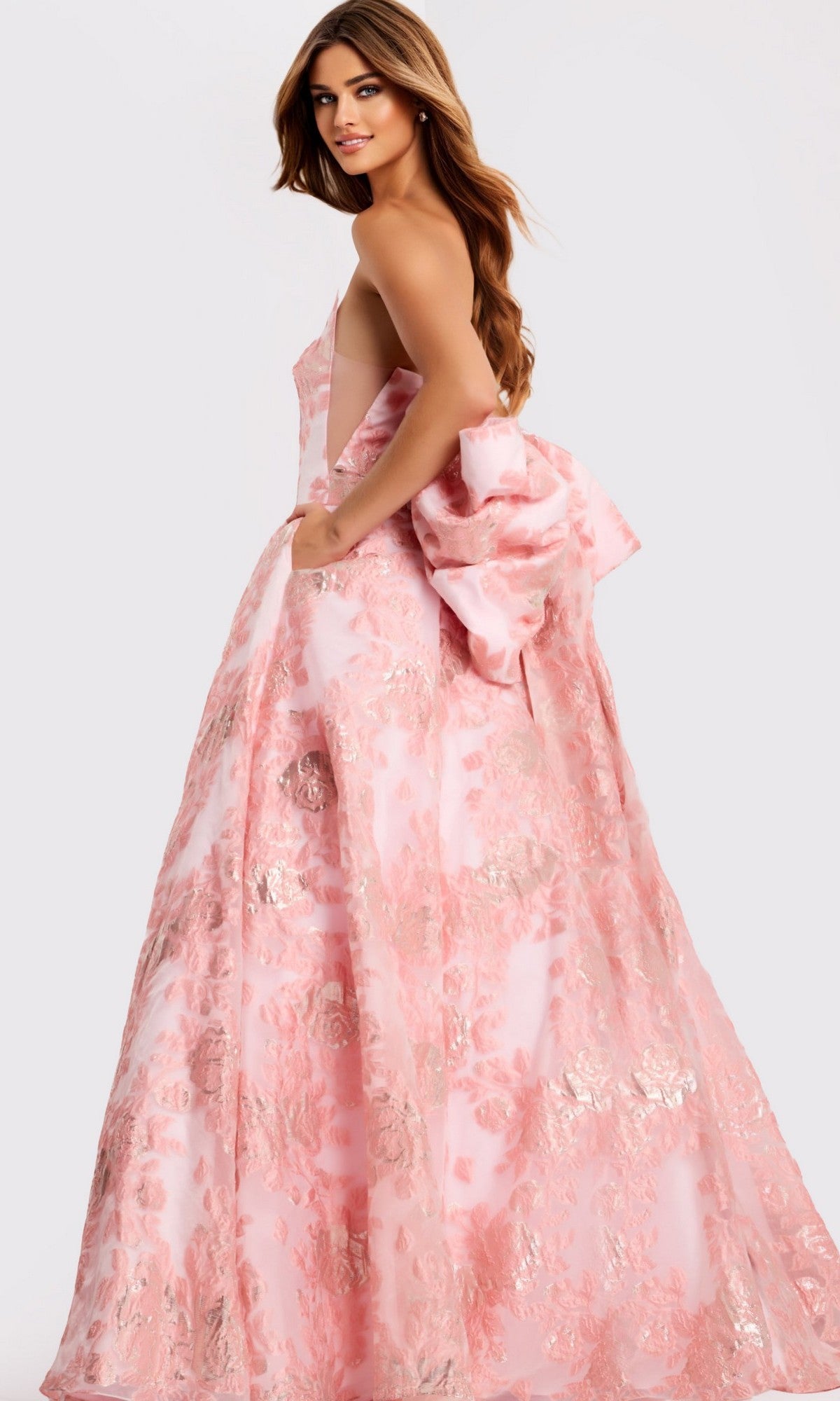 A woman looks over her shoulder in the Jovani 47702 long prom dress, a sleeveless pink gown with a floral pattern and a large bow at the back, set against a plain light background.